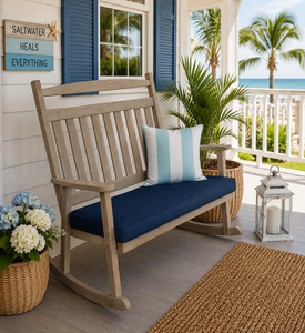 Weathered wood poly bench rocker with navy cushion on a coastal porch overlooking palm trees and the ocean.