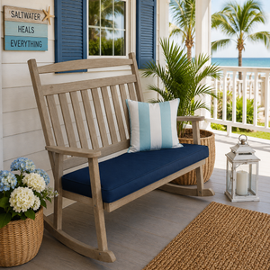 Weathered wood poly bench rocker with navy cushion on a coastal porch overlooking palm trees and the ocean.