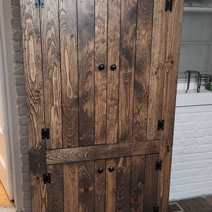 A rustic dark brown wooden storage cabinet with two upper doors and one lower door, featuring black metal hinges and knobs, set against a white brick wall.