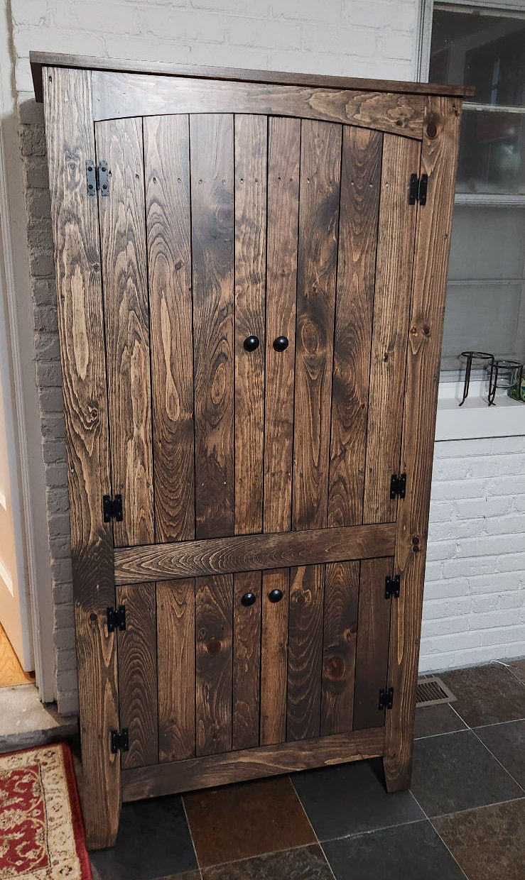 A rustic dark brown wooden storage cabinet with two upper doors and one lower door, featuring black metal hinges and knobs, set against a white brick wall.