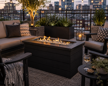 Black poly fire pit creates a cozy outdoor gathering space on a modern rooftop patio with city skyline views at dusk.
